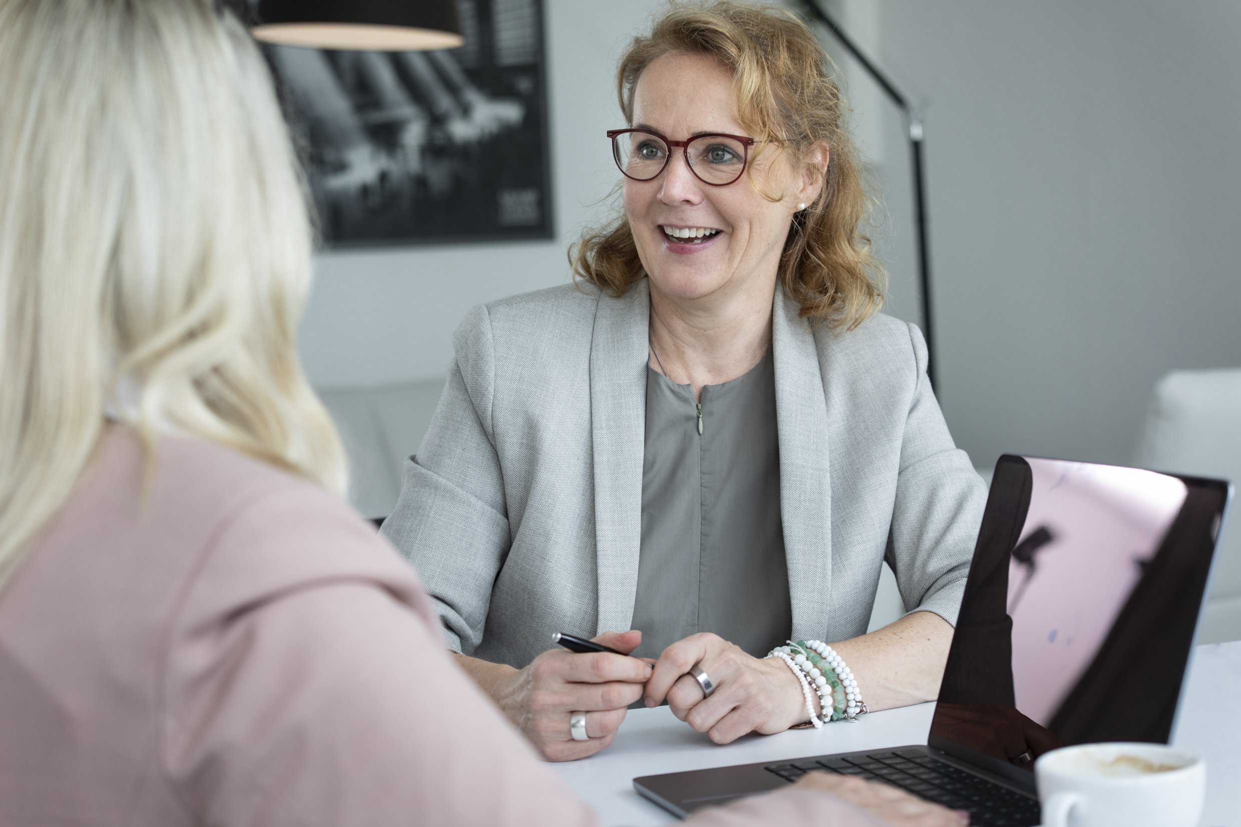 Zwei Personen sitzen nebeneinander am Tisch. Die blonde Frau hat einen Laptop und eine Tasse Kaffee vor sich. Die andere Frau, Maya Diekmann lächelt die andere Frau an.
