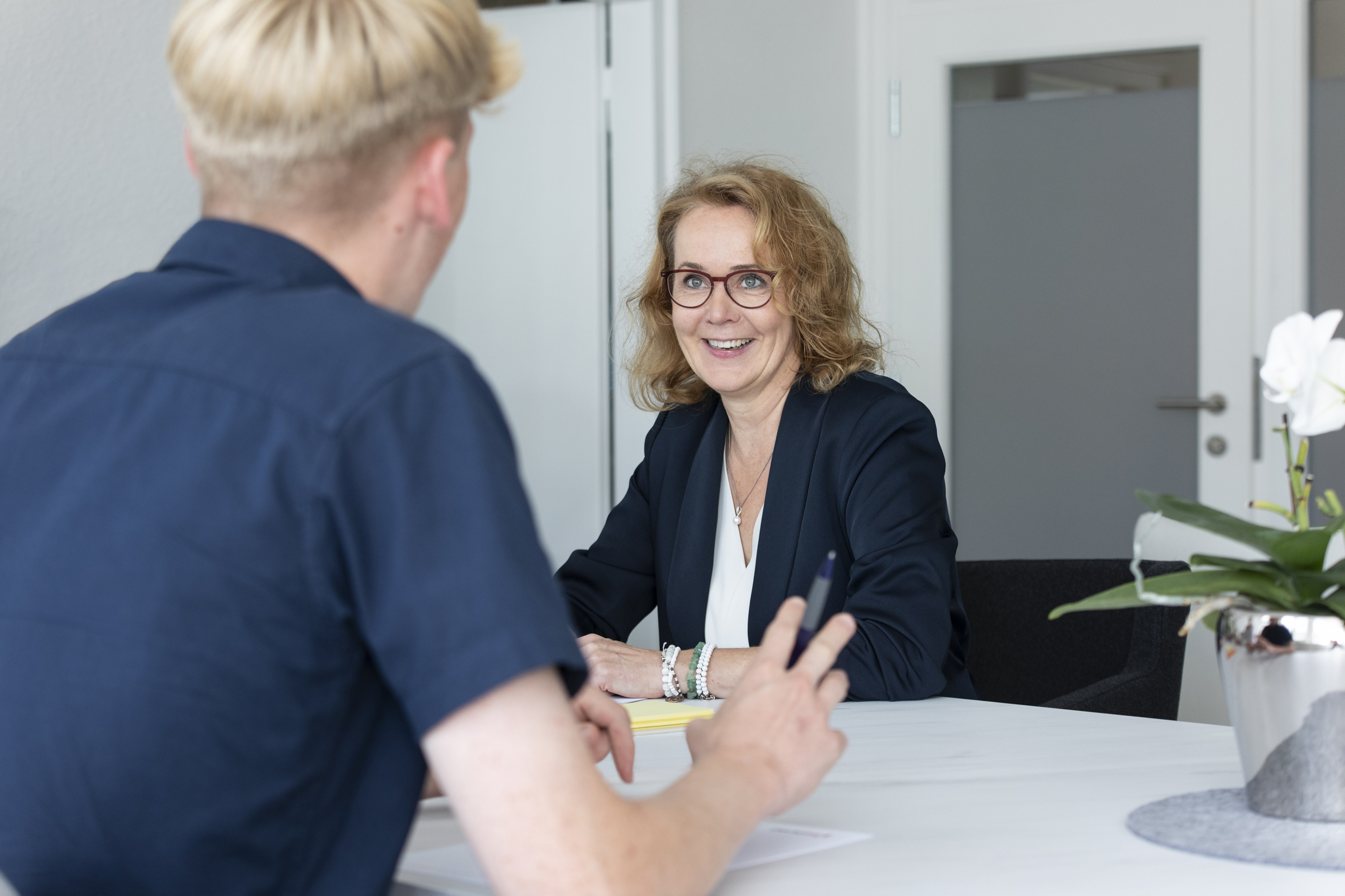 Eine blonde Frau mit Brille sitzt an einem weißen Tisch gegenüber von einem blonden Mann. Von dem Mann sieht man nur den Rücken und dass er mit seinen Händen einen Stift hochhält.