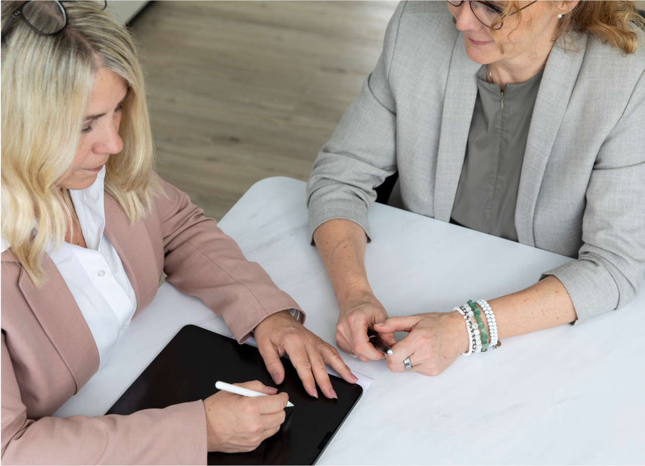 Zwei blonde Frauen mit jeweils einer Brille sitzen am Tisch. Die linke Frau hat einen Stift in der Hand und beschriftet etwas auf dem iPad.